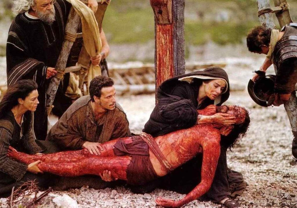 Fotograma de la película 'La Pasión de Cristo' mostrando el desprendimiento de Jesús de la Cruz, está en el suelo, su Maria lo sostiene por la cabeza acariciando su mano. dos apóstoles más le sujetan las piernas