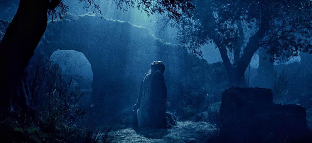 Jesús de Nazareth arrodillado en oración en el Huerto de Getsemaní, entre dos olivos, bajo la luz de la luna en una escena nocturna.