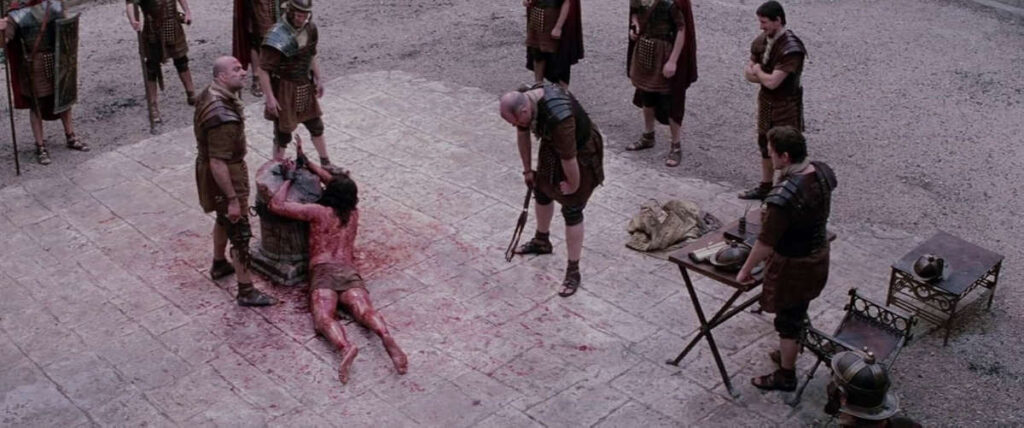 Fotograma de la película 'La Pasión de Cristo' mostrando la flagelación de Jesús de Nazaret, tumbado boca abajo y atado, con la espalda ensangrentada. Un romano descansa con el látigo, agotado, rodeado de soldados en una plaza.